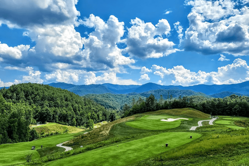 A vibrant green golf course spans the foreground with rolling hills and a blue sky dotted with fluffy clouds in the background.