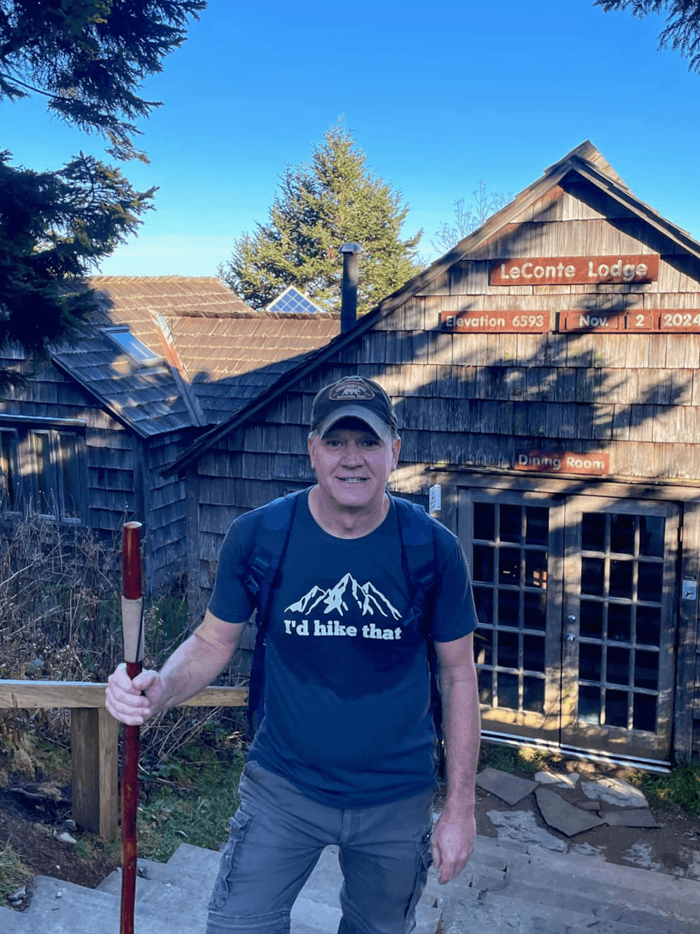 A man stands on steps outside LeConte Lodge, smiling while holding a hiking stick.