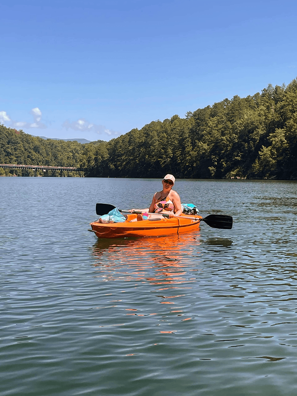 A person relaxes in an orange kayak on a calm lake surrounded by green trees and a clear blue sky.
