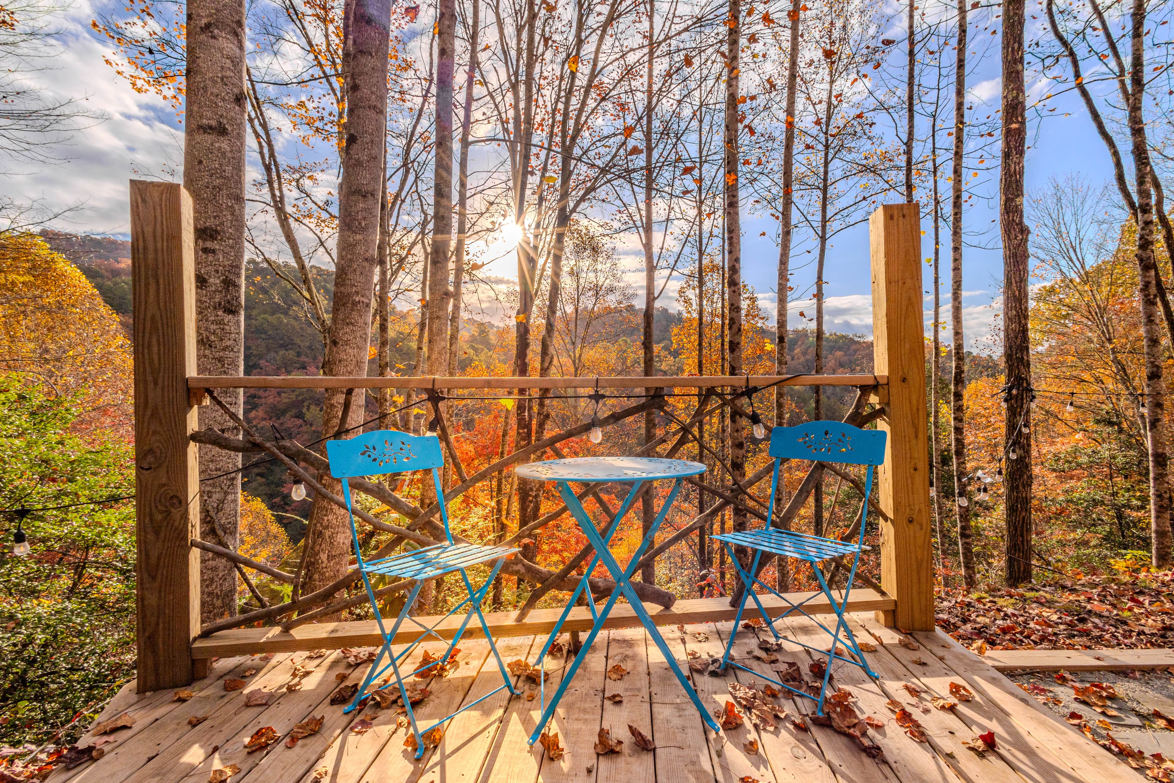 A blue bistro table and two chairs on a wooden deck overlook a forest of autumn trees.