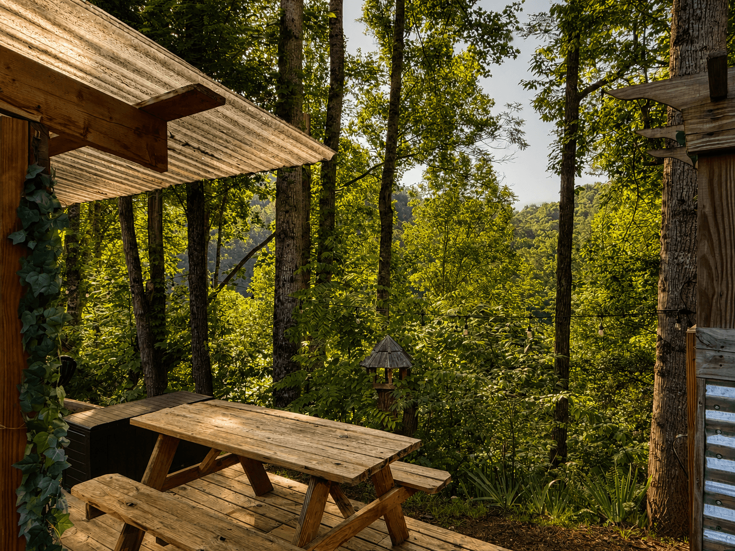 A wooden picnic table sits under a corrugated roof, surrounded by lush green trees.