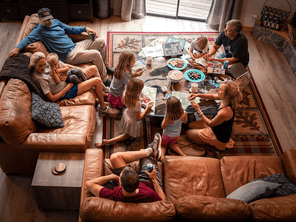 A multigenerational family gathers around a coffee table filled with snacks in a cozy living room.