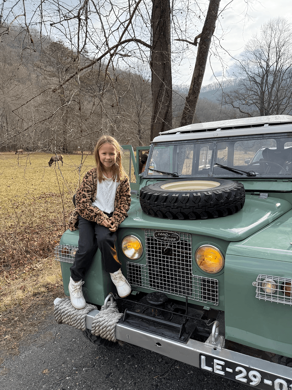 A girl sits happily on the front of a vintage green Land Rover parked in a rustic landscape.