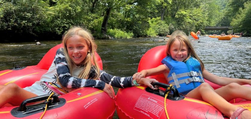 Two girls in colorful inner tubes smile and hold hands while floating on a river surrounded by trees.