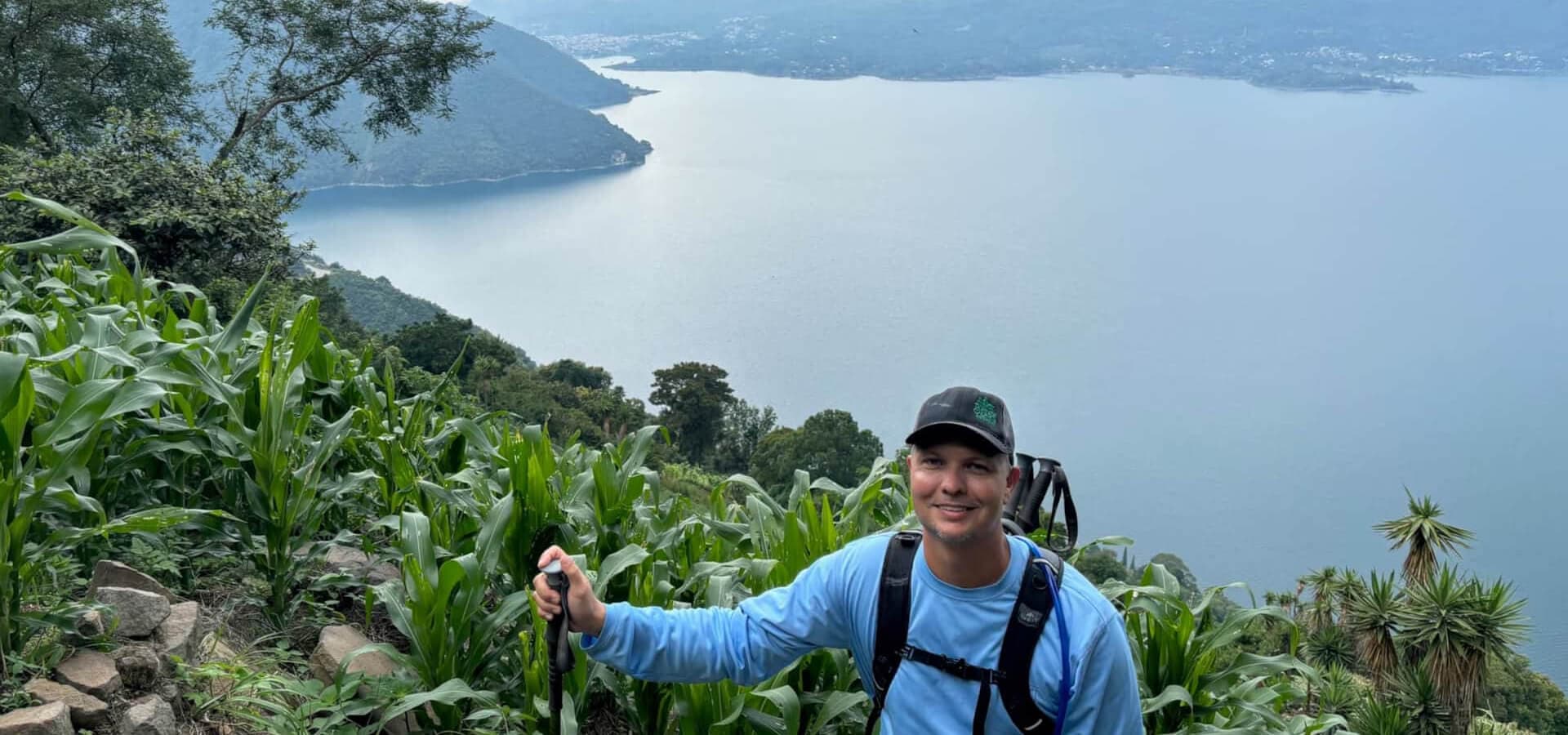 A hiker smiles while standing among green vegetation with a scenic lake in the background.