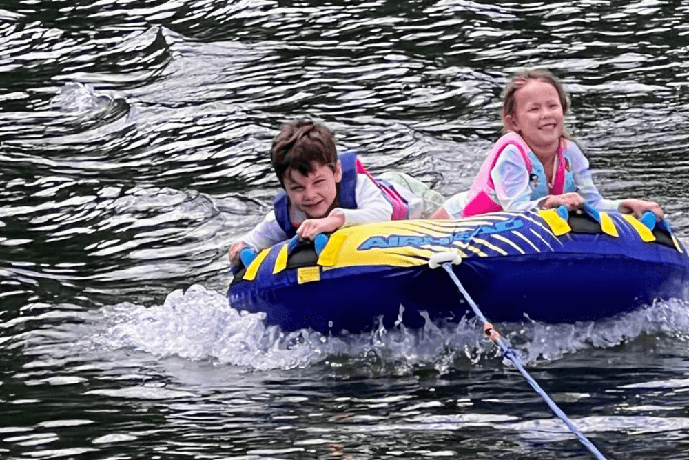 Two children joyfully ride a blue and yellow inflatable tube being towed on water.