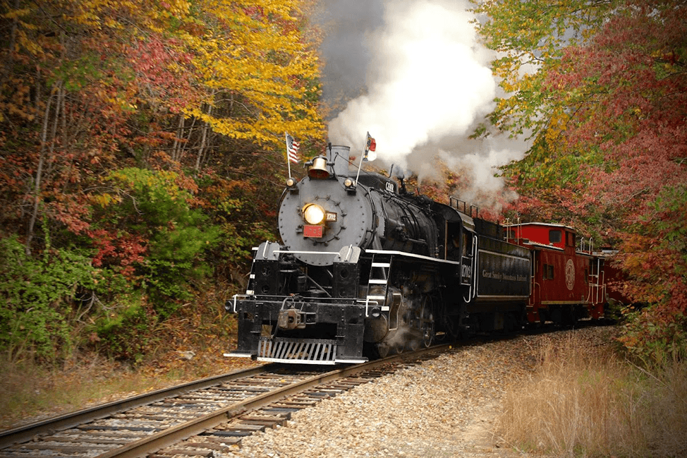 A steam locomotive chugs along a track surrounded by vibrant autumn foliage.