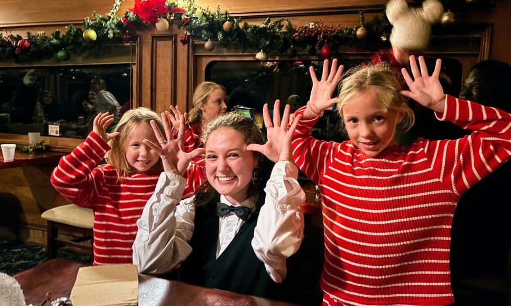 A smiling young woman in a tuxedo poses playfully with two girls in matching red-striped pajamas, all making hand gestures aboard the Polar Express train.