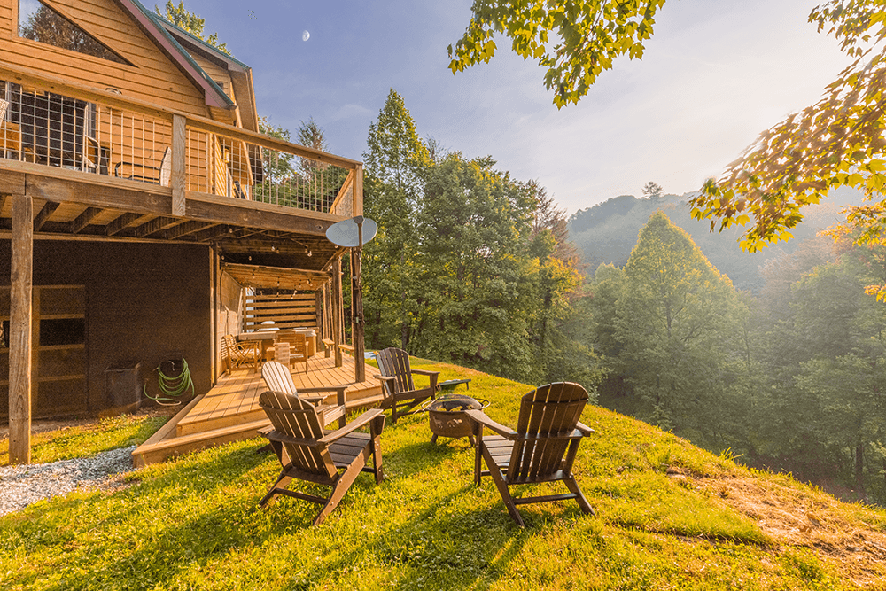 A cozy wooden cabin with a deck overlooks a green hillside, featuring Adirondack chairs arranged in front.