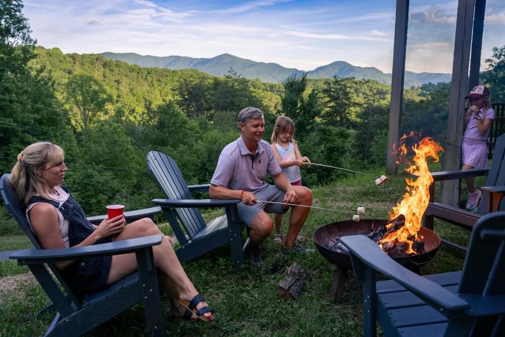A family enjoys a cozy evening by a fire pit with a scenic mountain backdrop.