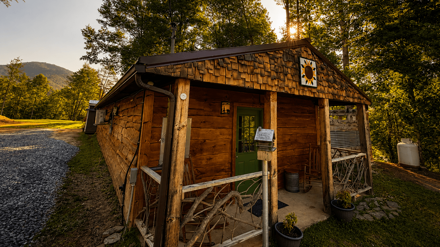 A rustic wooden cabin with a green door and a sun symbol, surrounded by trees and a gravel path.