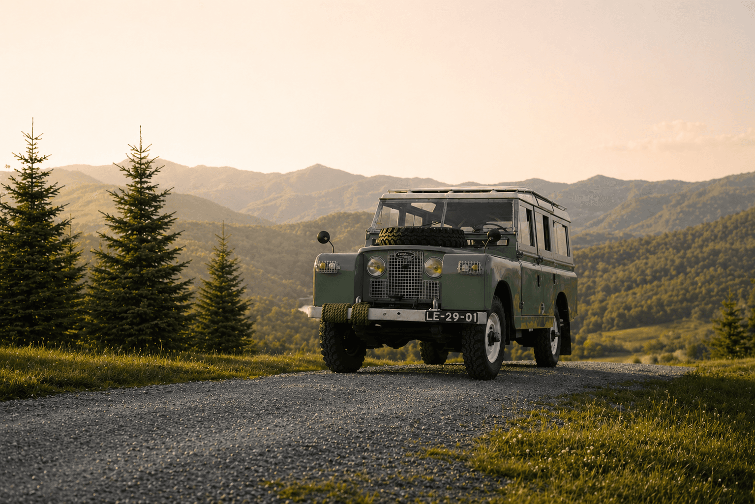 A vintage green Land Rover approaches on a gravel road surrounded by mountains and evergreen trees.