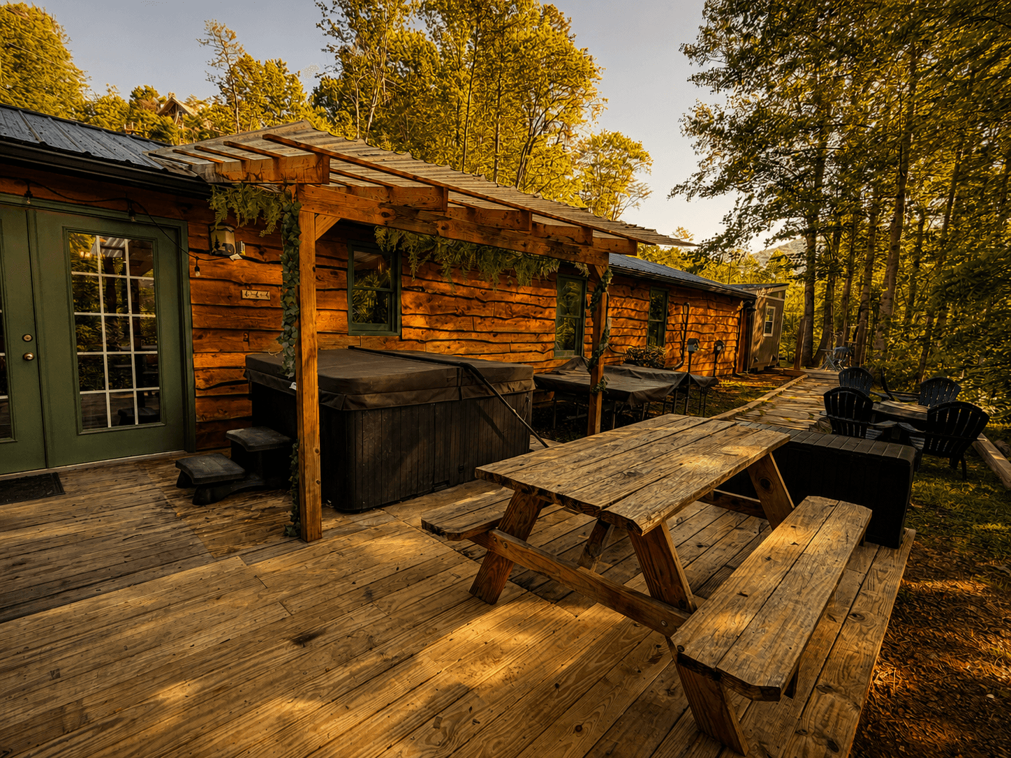 A rustic wooden cabin exterior features a deck with a picnic table, hot tub, and seating area surrounded by trees.