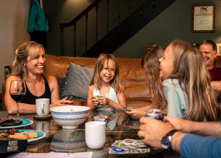 A group of children and adults laugh and play cards around a coffee table in a cozy living room. A group of children and adults laugh and play cards around a coffee table in a cozy living room.