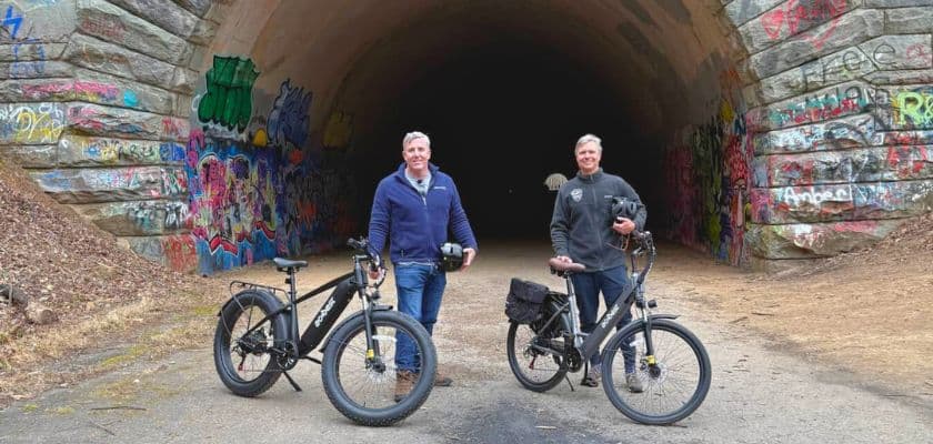 Two men stand with their bicycles in front of a graffiti-covered tunnel entrance.