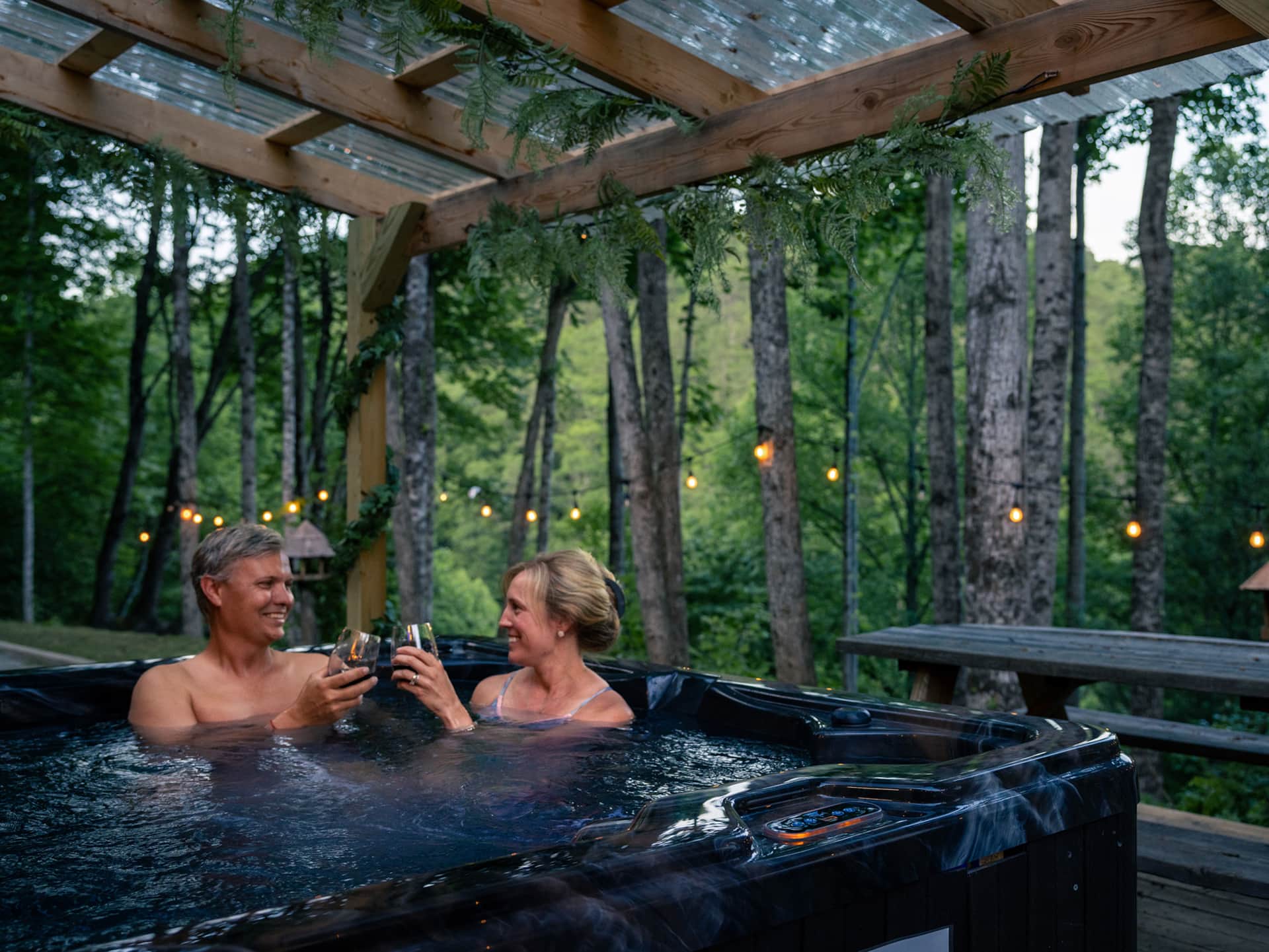 A man and woman enjoy drinks together in a hot tub surrounded by trees and soft lighting.