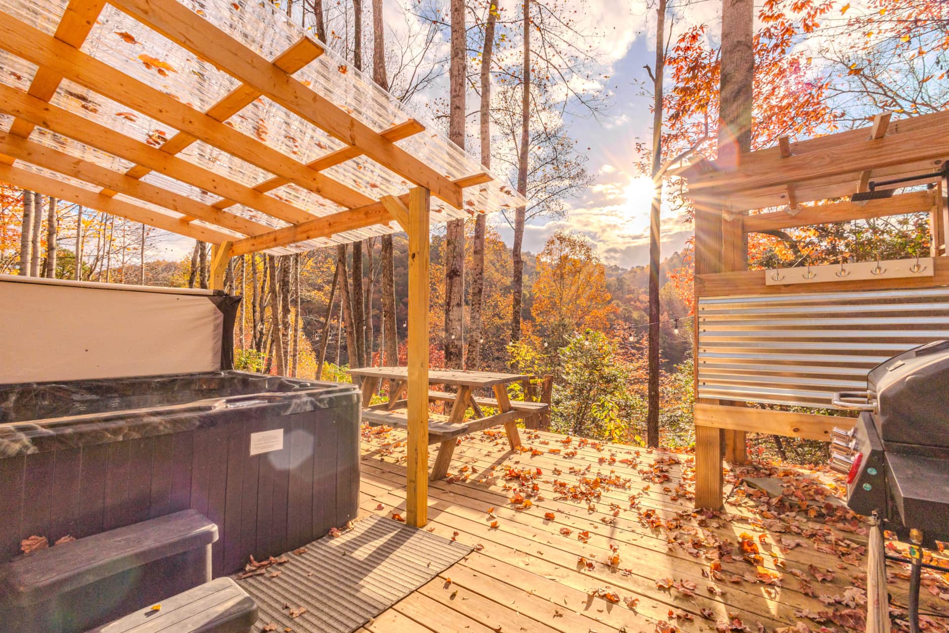 A cozy outdoor deck with a hot tub, picnic table, and grill, surrounded by autumn-colored trees and a sunset.