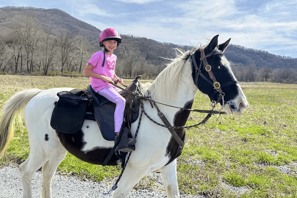 A girl in a pink helmet rides a black and white horse through a grassy field.