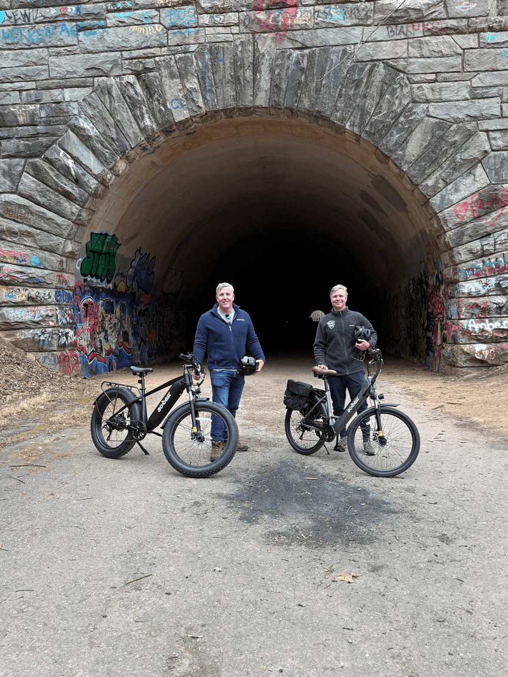 Two men standing with bicycles in front of a graffiti-covered tunnel.
