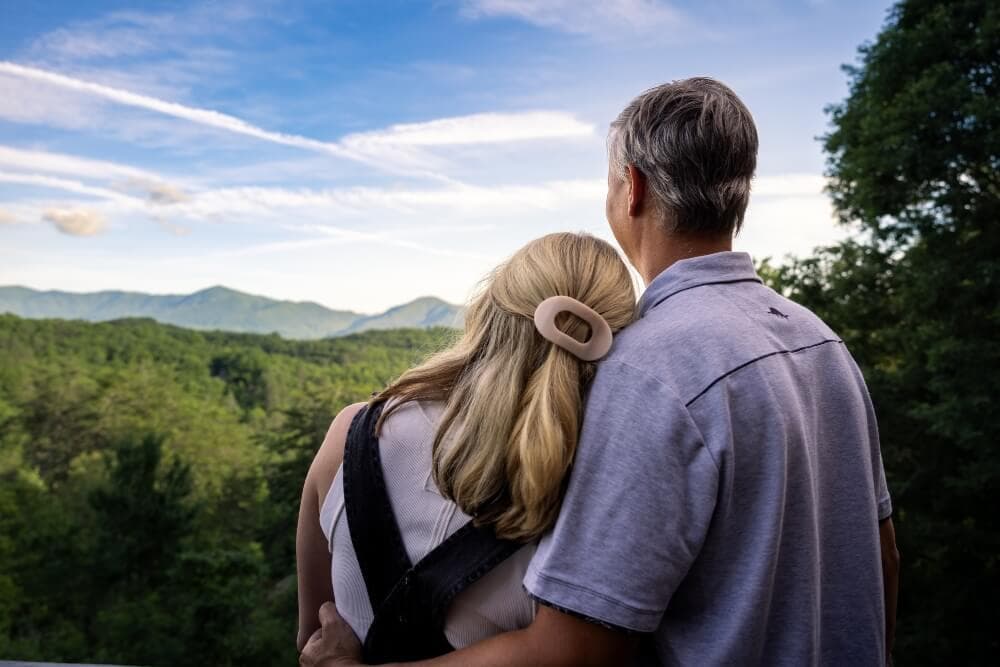 A couple stands together, embracing while gazing at a scenic mountain view under a blue sky.