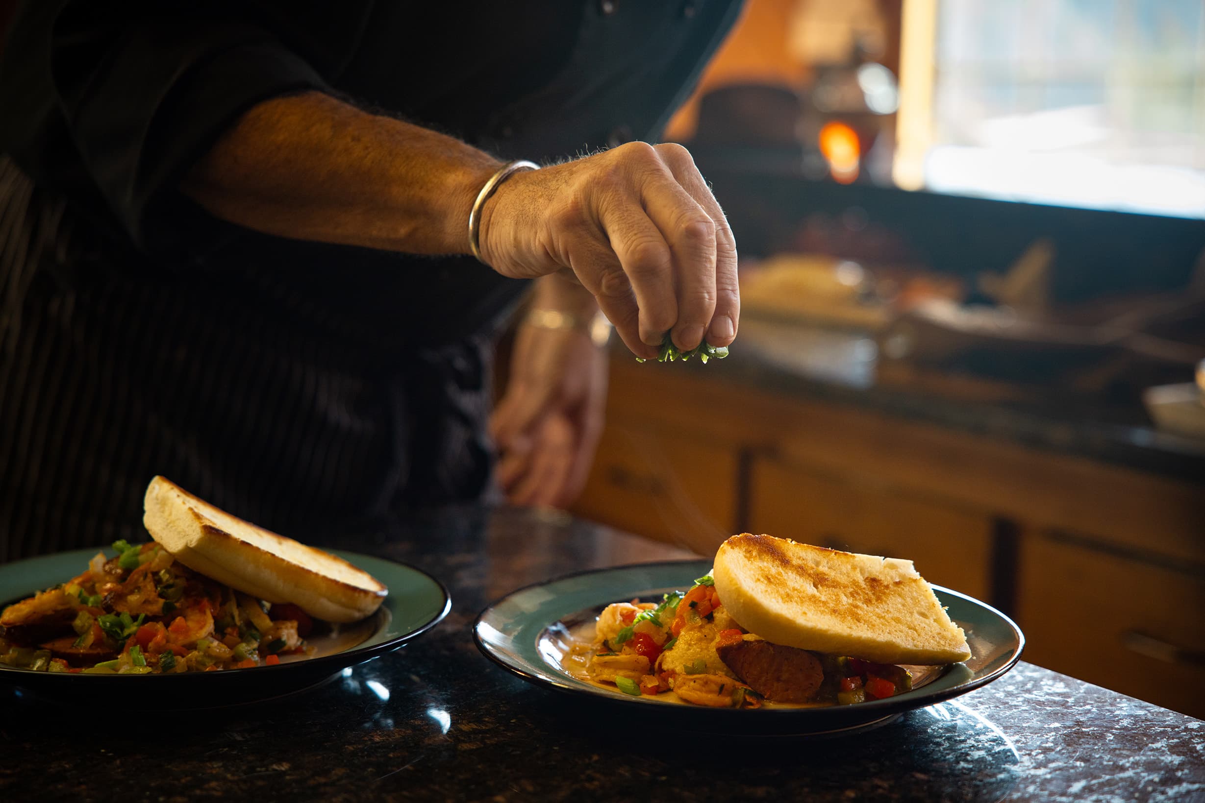 A chef sprinkles herbs over two plates of colorful dish with toast.