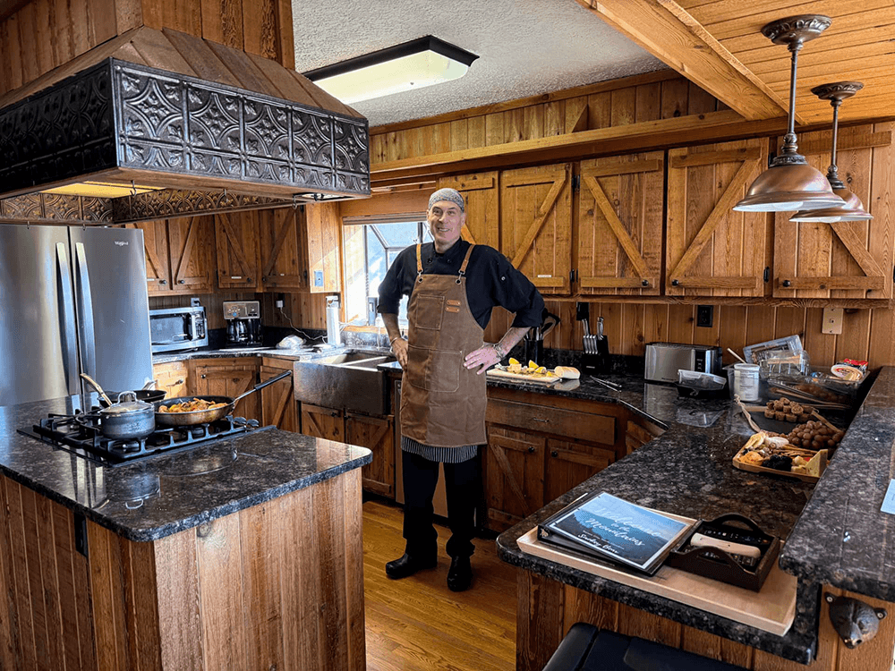 A chef stands in a rustic kitchen with wooden cabinets, preparing food.