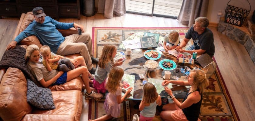 An overhead view of adults and children gathered around a coffee table playing card and board games in a cabin living room.
